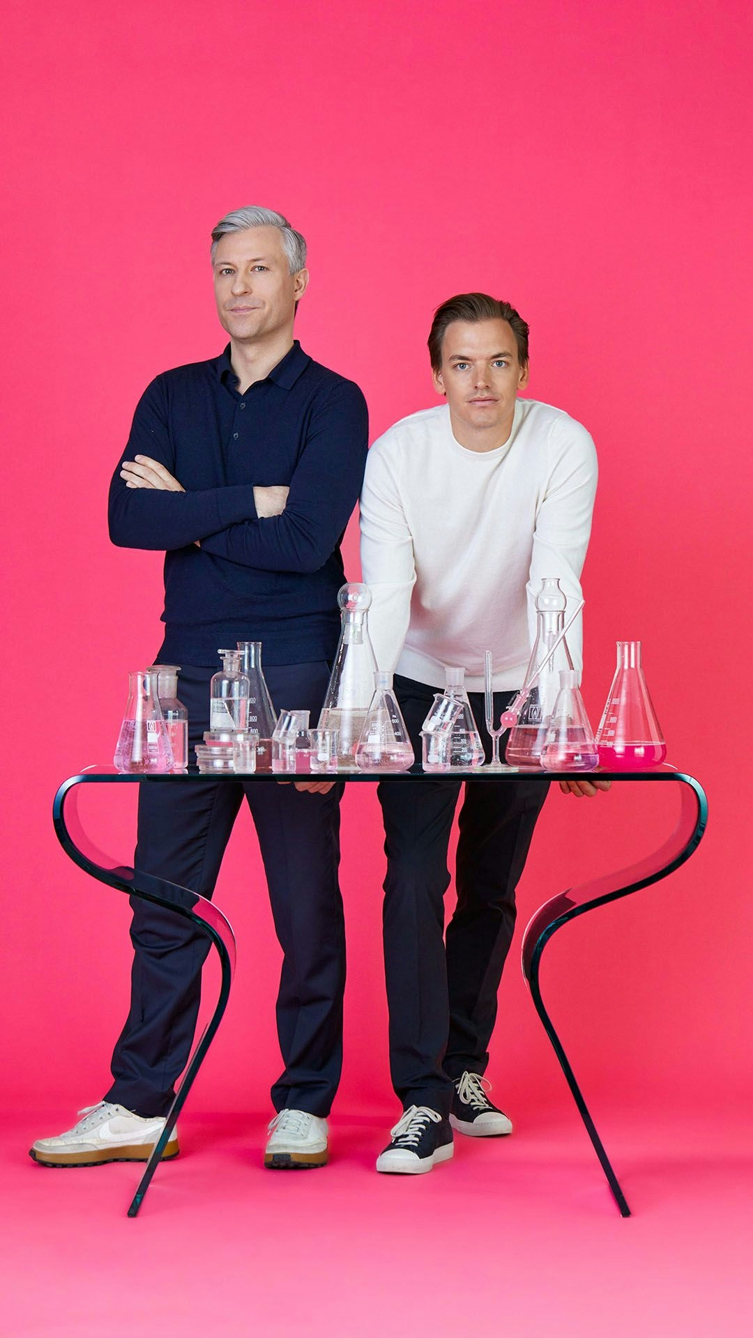 Two men standing behind a table full of beakers in front of a pink background